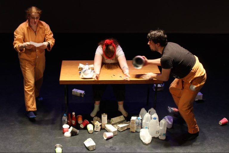 A shot of three people on stage. One stands to the left in an orange boiler suit. To their left another is slumped over a table. To the right of the table another quickly moves a metal object. 