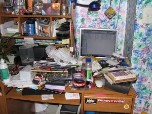 Photograph of a messy desk. The computer is barely visible amongst papers and junk.
