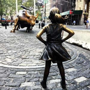 Photograph of the Fearless Girl statue in New York. The statue is of a young girl, standing with her hands on her hips, facing down a statue of an enormous bull.