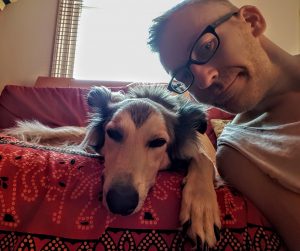 Photograph of the website author and his dog. His dog lies on the sofa gazing sleepily at the camera. Richard leans in from the side.