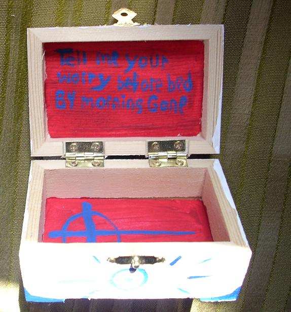 Photo of a handmade wooden worry box, Painted on the inner lid of the open box in blue paint are the words "Tell me your worry before bed. By morning gone". The interior of the box is red and a blue cross is painted on the inner bottom of the box.