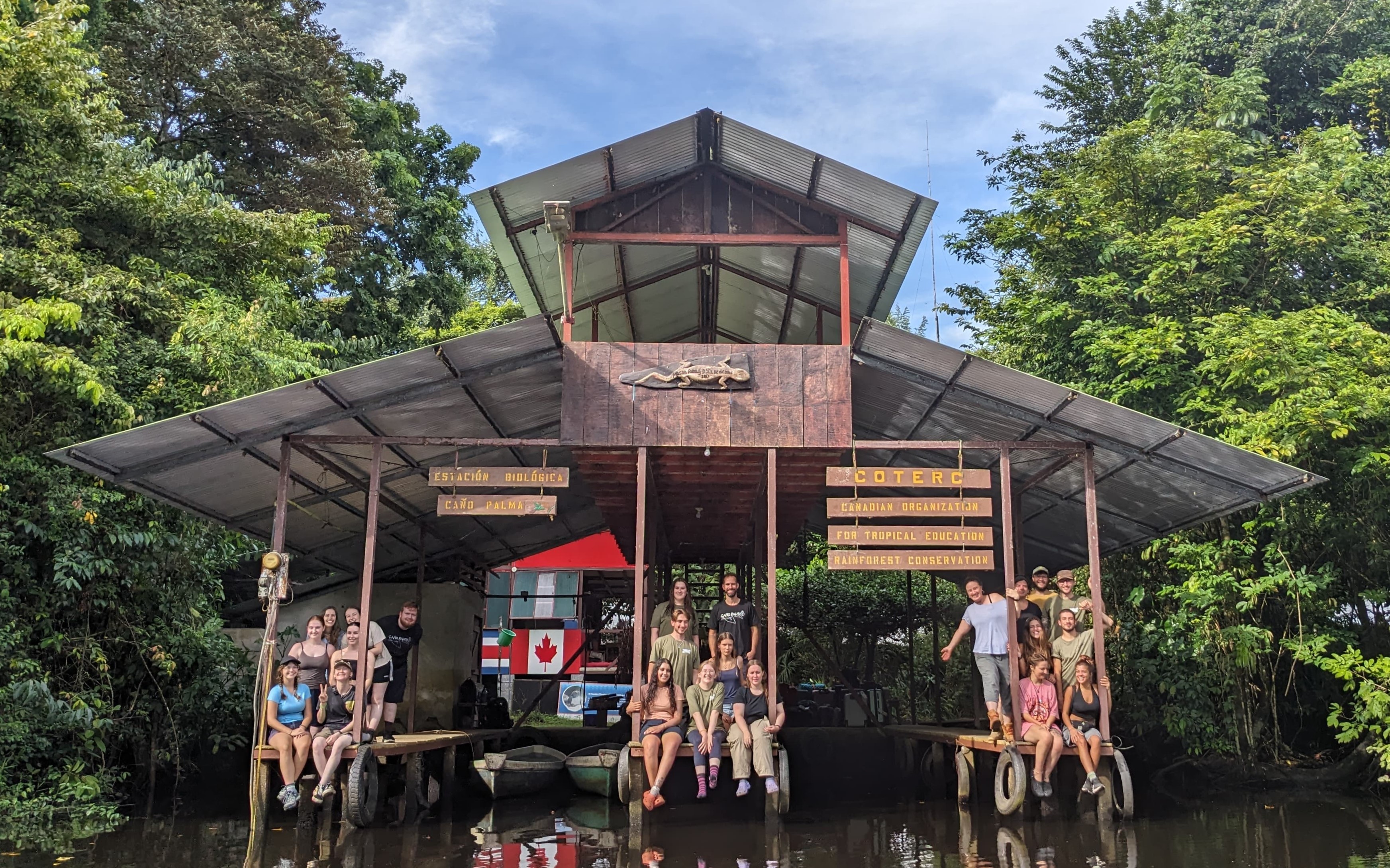 A group of people sit on the edge of a wooden station, built on the bank of a river. They smile at the camera, some with their feet dangling above the water as they sit on the edge. Some people are waving.