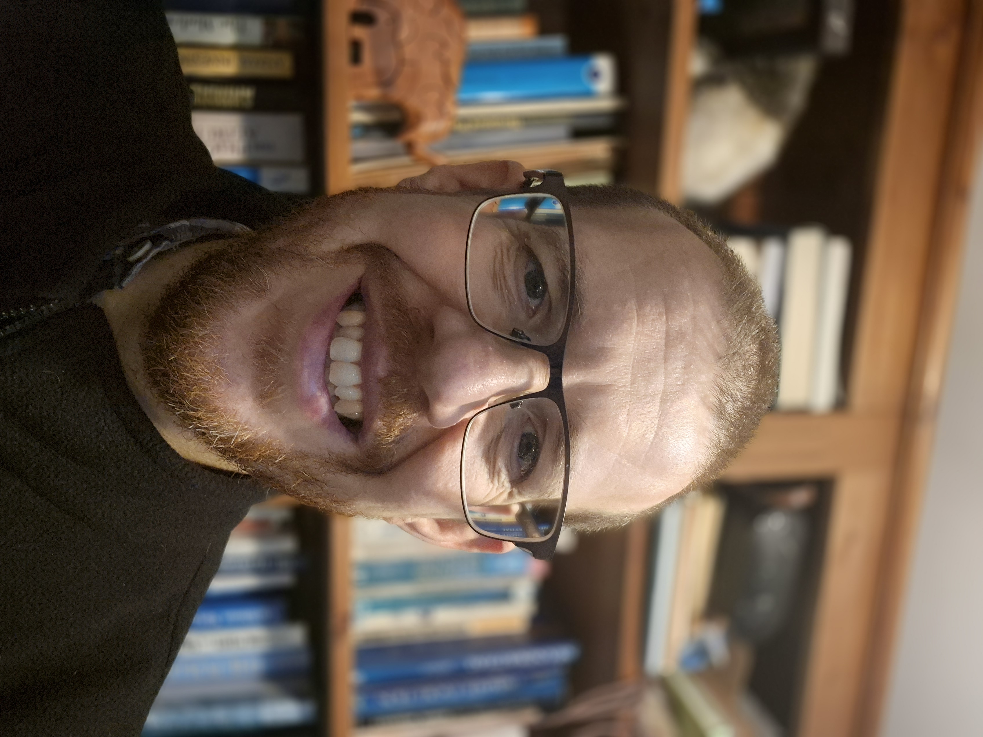 A man smiles at the camera. In the background are bookshelves.