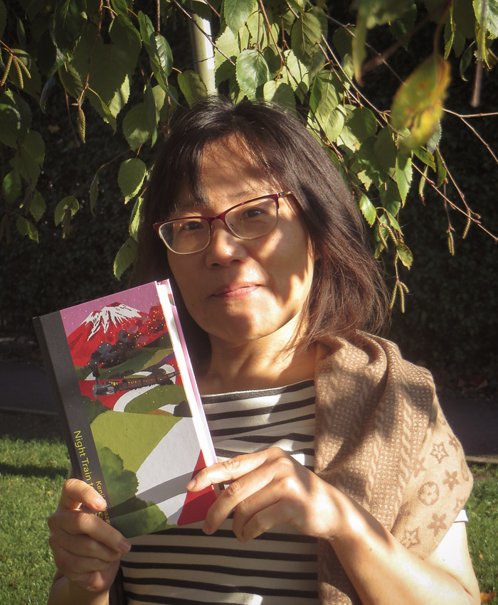 A woman stands in dappled shade under a tree, holding a colourful book