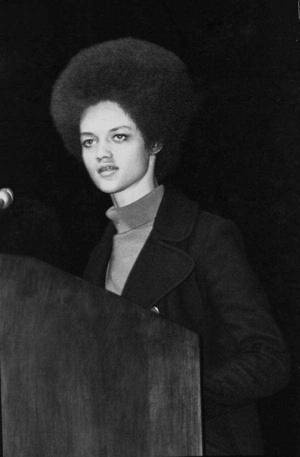 A woman with an afro stands at a lecturn mid-speech