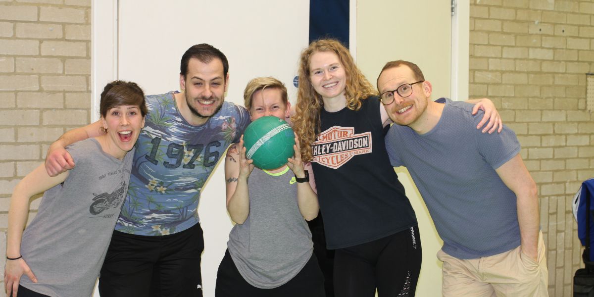 Five team members posing for a photo with arms around each others shoulders, smiling. The player in the middle is holding a dodgeball up to their face to hide from the camera. 