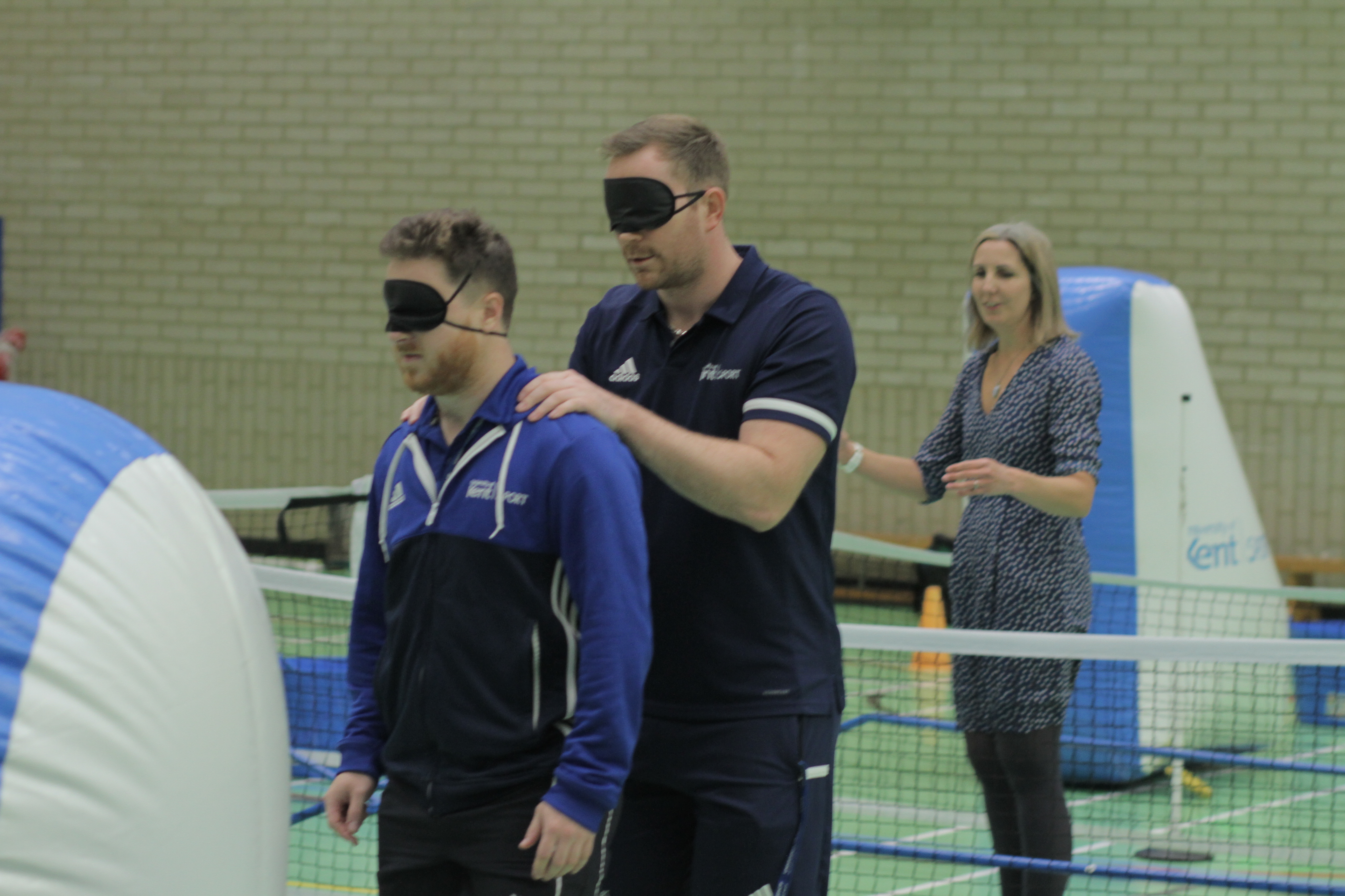 Two staff members stood one in front of the other. The person at the back has his hands rested on his partners shoulders. They are both wearing blindfolds.