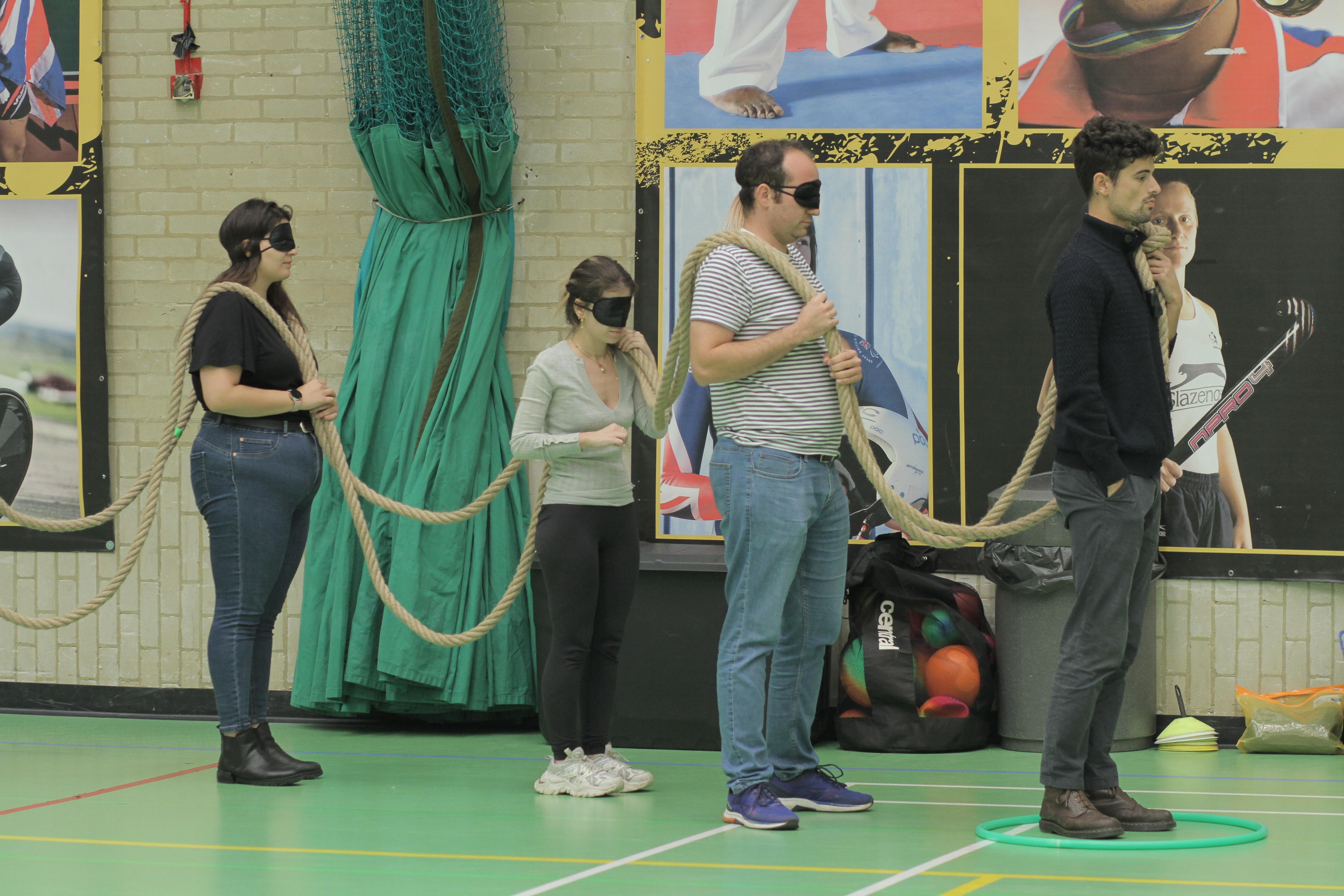 Four people standing in a line, three with blindfolds on, holding a rope, slung over their shoulders. 