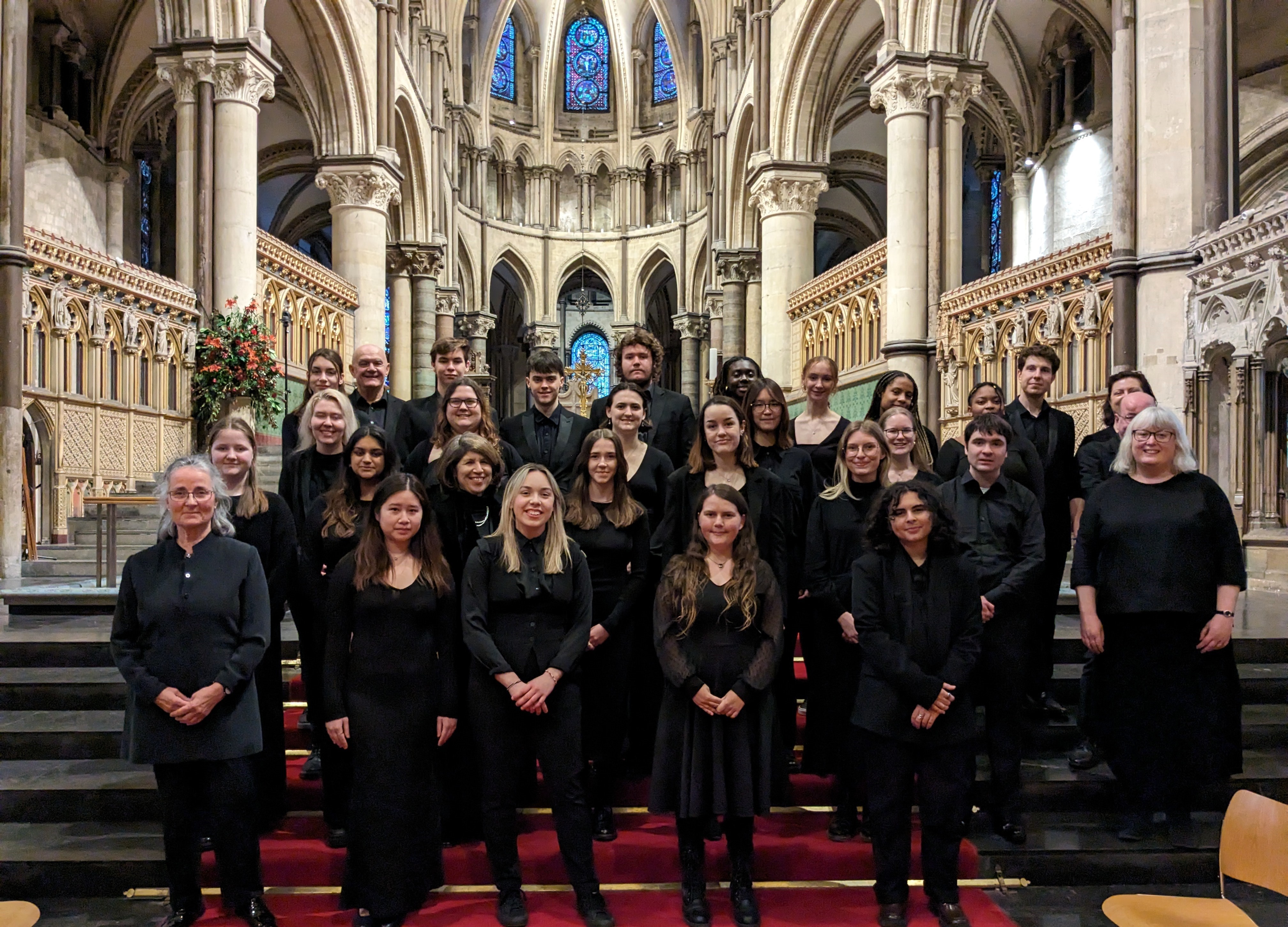 Group of thirty singers dressed in formal black standing on red-carpeted steps amidst the ornate architecture of Canterbury Catrhedral