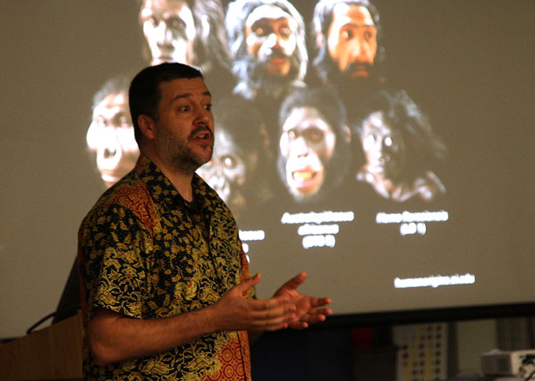 Dr Tocheri in front of a slide of different hominin likenesses across the various evolutionary stages of early man