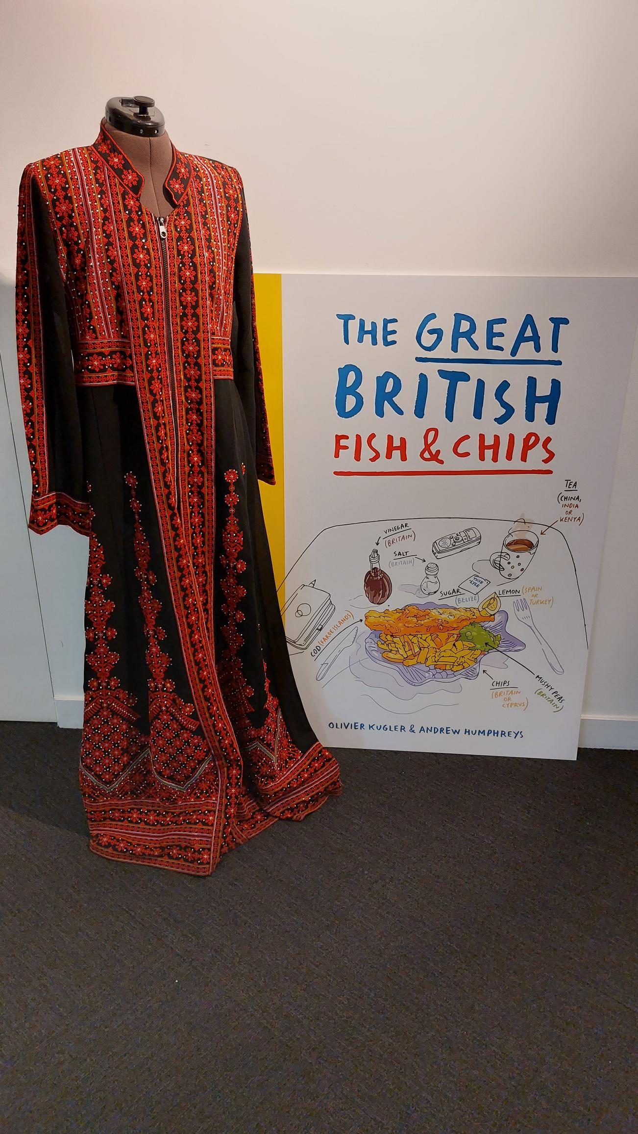 Black, Red and White handstitched dress in traditional Palestinian style next to an exhibition board for the Great British Fish and Chips exhibition. The dress is exhibited in the exhibition. 