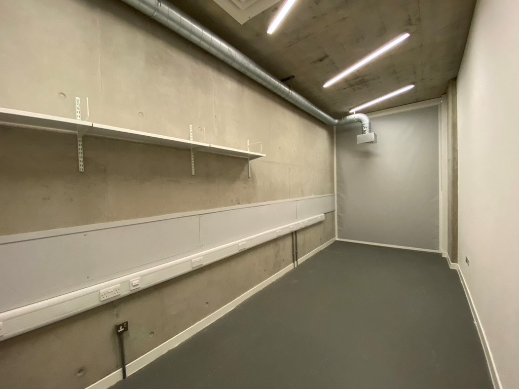 A photograph of an empty room, with concrete walls , with white shelves high up on the wall.