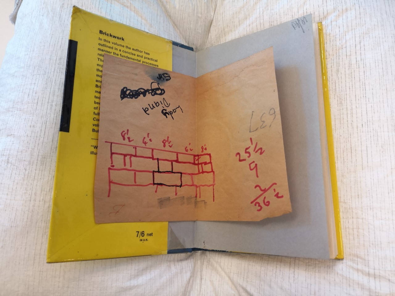 A photograph of a book open to the front page. Inserted in the book is a piece of paper with an image of a brick wall drawn in red pen, alongide some measurements. 