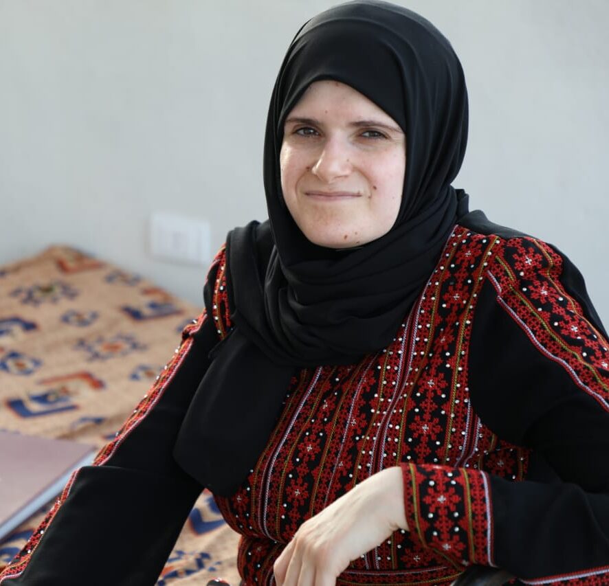Portrait image of Basma El Doukhi - wearing a black headscarf and a black, red, white and gold decorated dress 