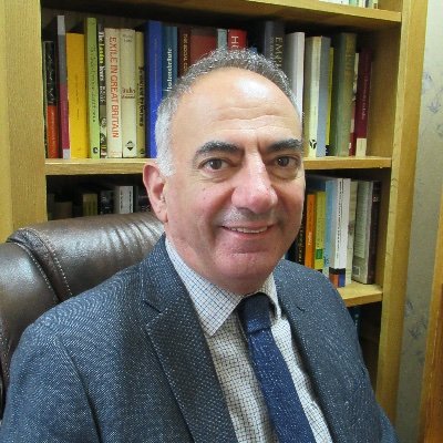 Portrait image, head and shoulders, of Professor Panikos Panayi, looking directly at the camera and wearing a blue suit and tie with a bookcase in the background