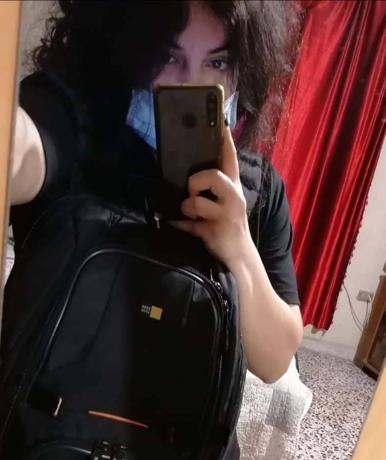 Portrait image of Rania Saadallah showing her holding her phone taking a selfie, wearing a bag on her front and wearing a face mask