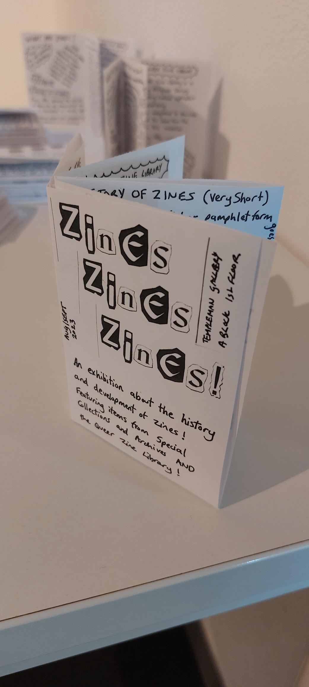 Small black and white zine, standing up with the front cover visible showing the text "Zines Zines Zines! Templeman gallery A Block 1st Floor. Aug/Sept 2023. An exhibition about the history and development of zines! Featuring items from Special Collections and Archives AND the Queer Zine Library!" In the background of the image in soft focus are other copies of the zine displayed on the table. 