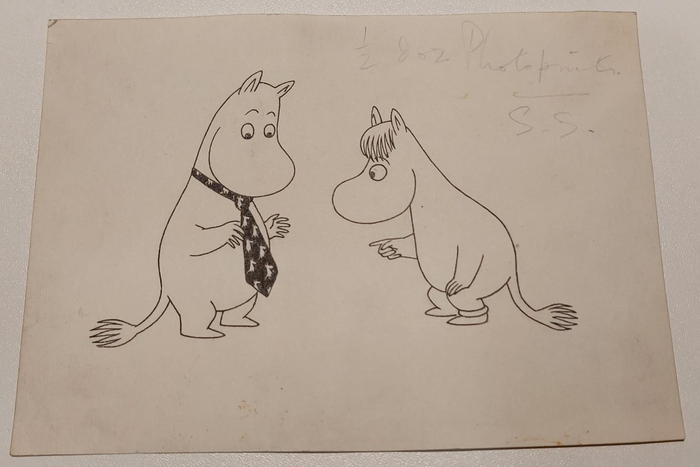 Drawing of two moomins by Tove Janssen. Moomintroll is standing wearing a neck tie with moomins on it, facing Snorkmaiden who is looking and pointing at the tie. 