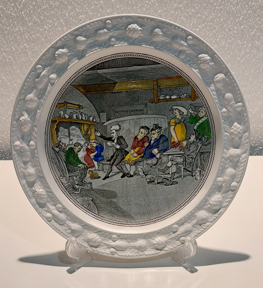 Ceramic plate on a plate stand showing an image of Dr Syntax, a character created by Thomas Rowlandson as a satire of William Gilpin, who was a supporter of the Picturesque movement. In the image Dr Syntax is reading his book to others in a tavern and they look bored and even asleep. 