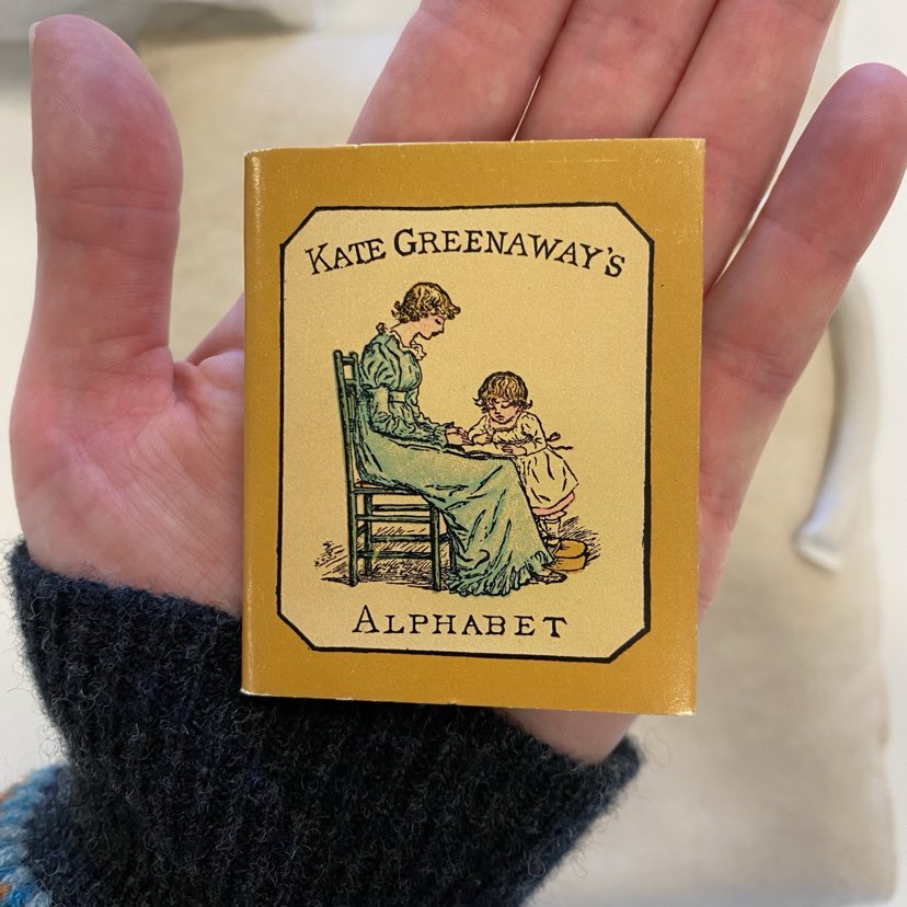 Image of the front cover of Kate Greenaway's alphabet, showing an illustration of a mother and child engaged in reading together. The book sits in the palm of a hand and measures 7cm.