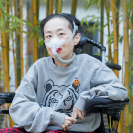 Photo of Alice Wong, an Asian American disabled woman with a mask over her nose attached to a tube for her ventilator. She is in a power wheelchair and wearing a gray sweatshirt with a tiger and leopard-print red and black pants. Behind her are bamboo tree.