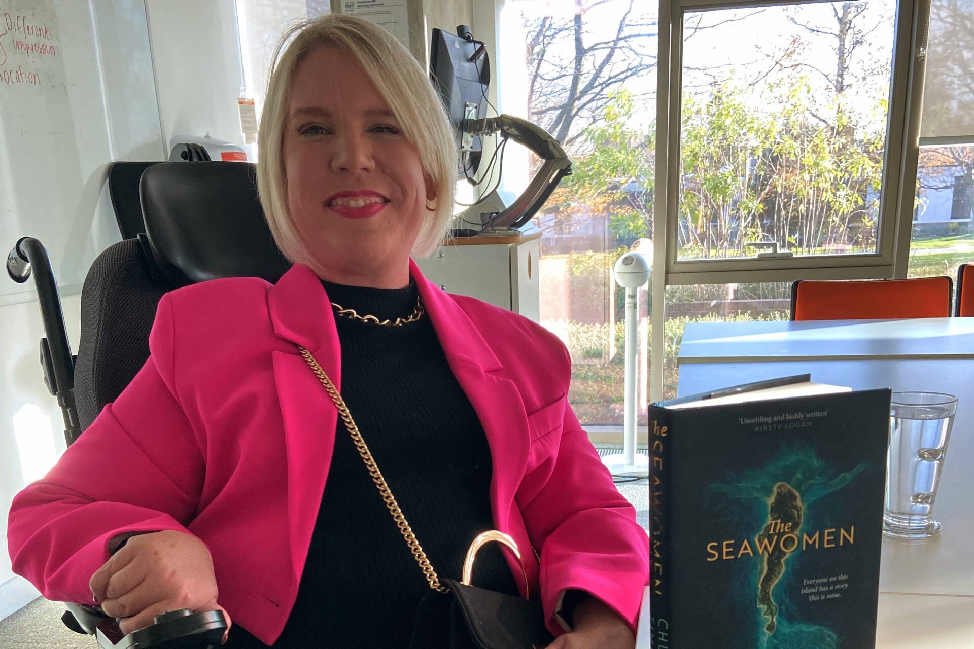 Chloe Timms with her book. Woman seated in wheelchair, with blonde hair and pink jacked. Book titled 'The Seawoman' on the table in front of her.
