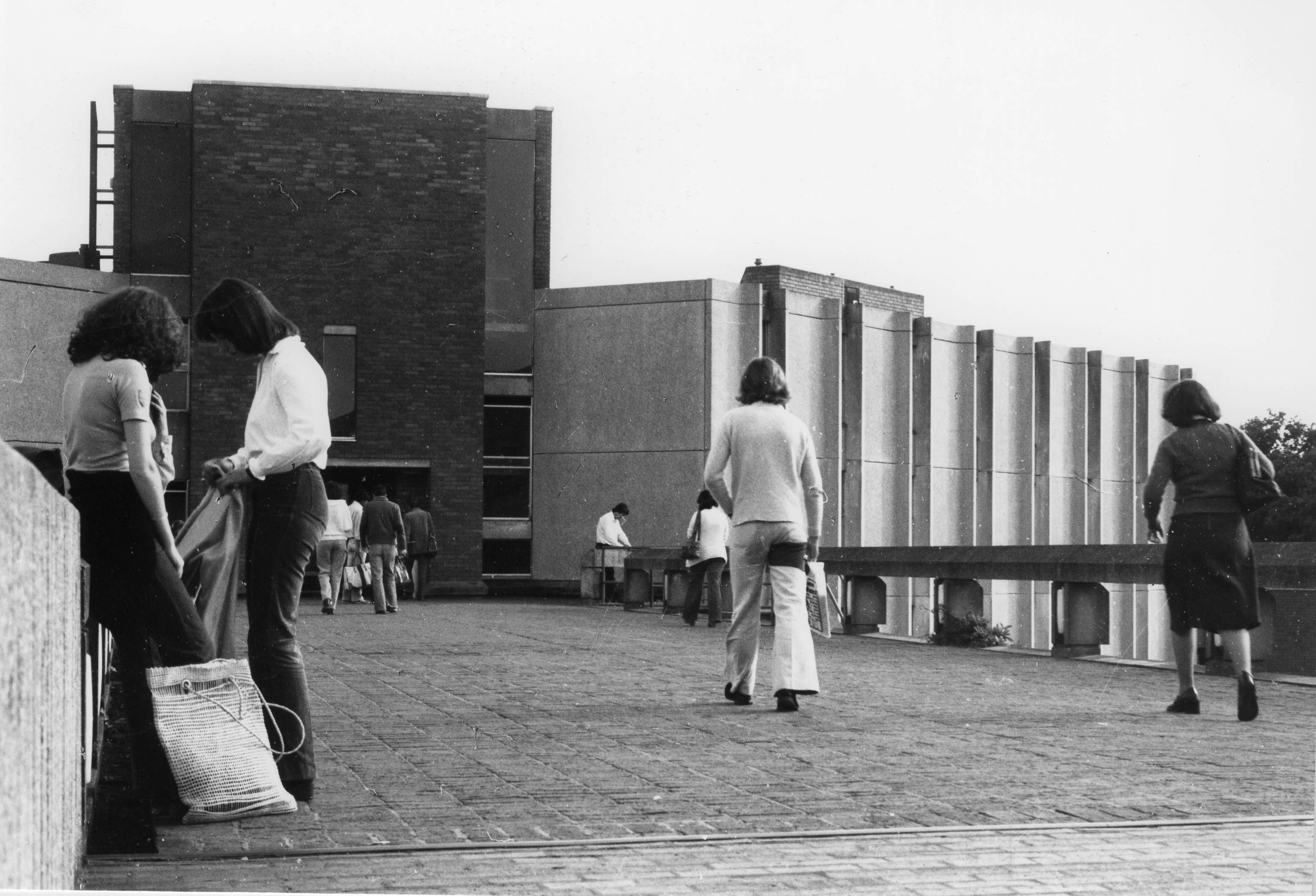 Black and white photograph of students on the causeway to the entrance of Eliot College, c1965. Two students are standing in a group on the left of the photo and two other students are walking towards the Eliot building in the background, with their backs to the camera