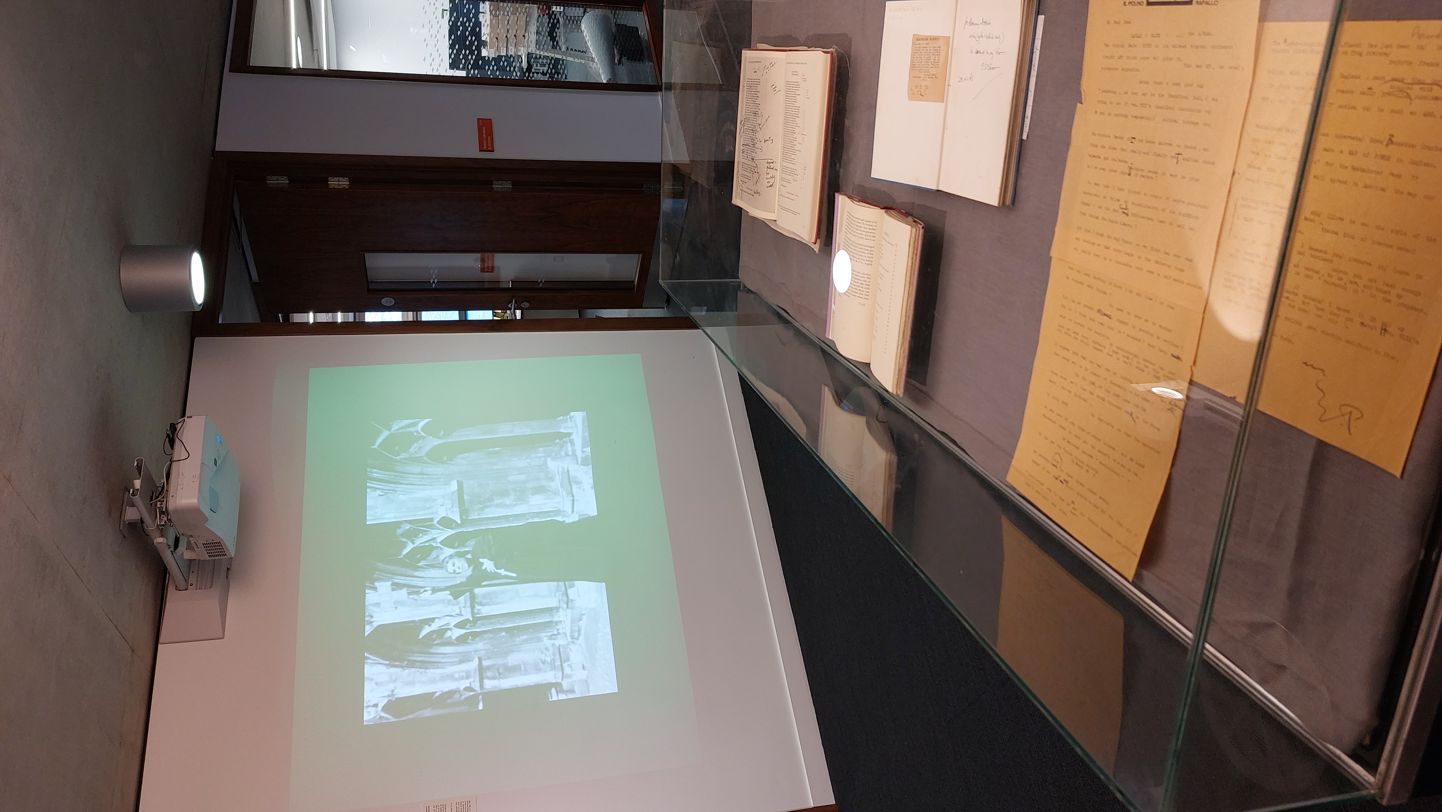 Image of a exhibition case in the foreground containing books and letters, and a film screen in the background showing a black and white image of a man in a cathedral setting 