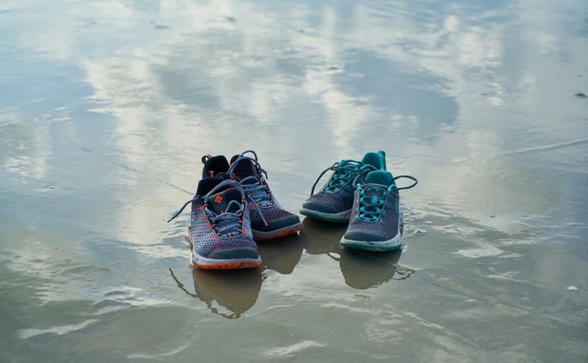 Photograph of two pairs of shoes on a sandy beach. Their reflection is captured in the layer of water on the sand.