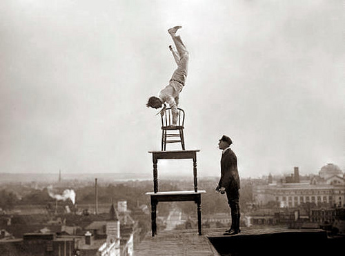 Photograph of a man performing a handstand on a wooden chair, which is itself balanced on two wooden tables on the edge of a tall building. Another man stands nearby.