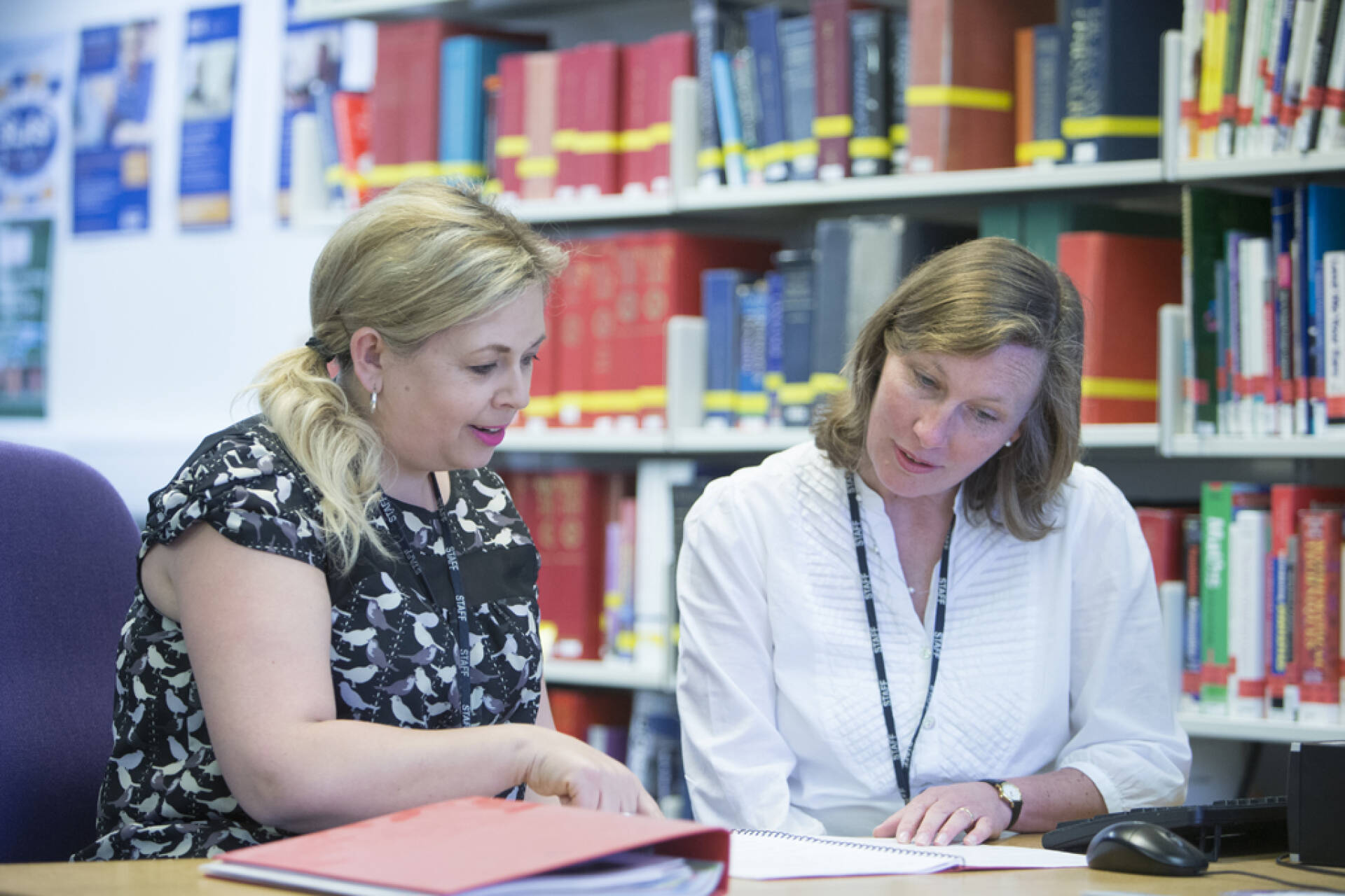 Two female colleagues studying notes in a notebook with books on library shelves in background