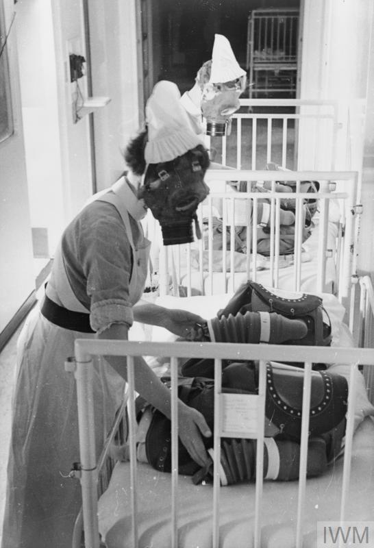 The black and white photo shows nurses in uniform wearing gas masks. They are fitting large gas masks on the babies in their care in a crib