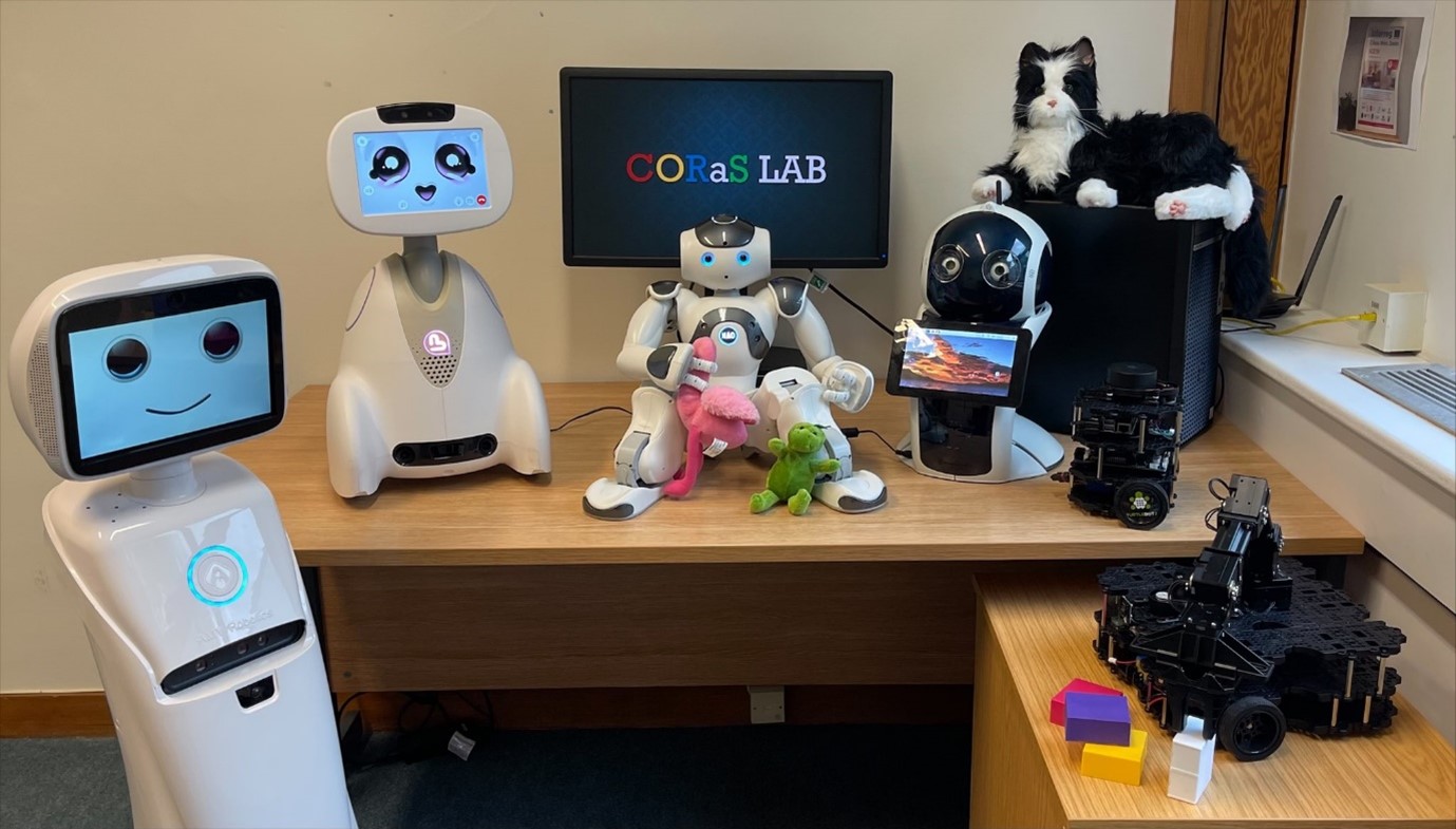 A computer monitor on a desk, Coras Lab written across the screen, is surrounded by robots and soft toys