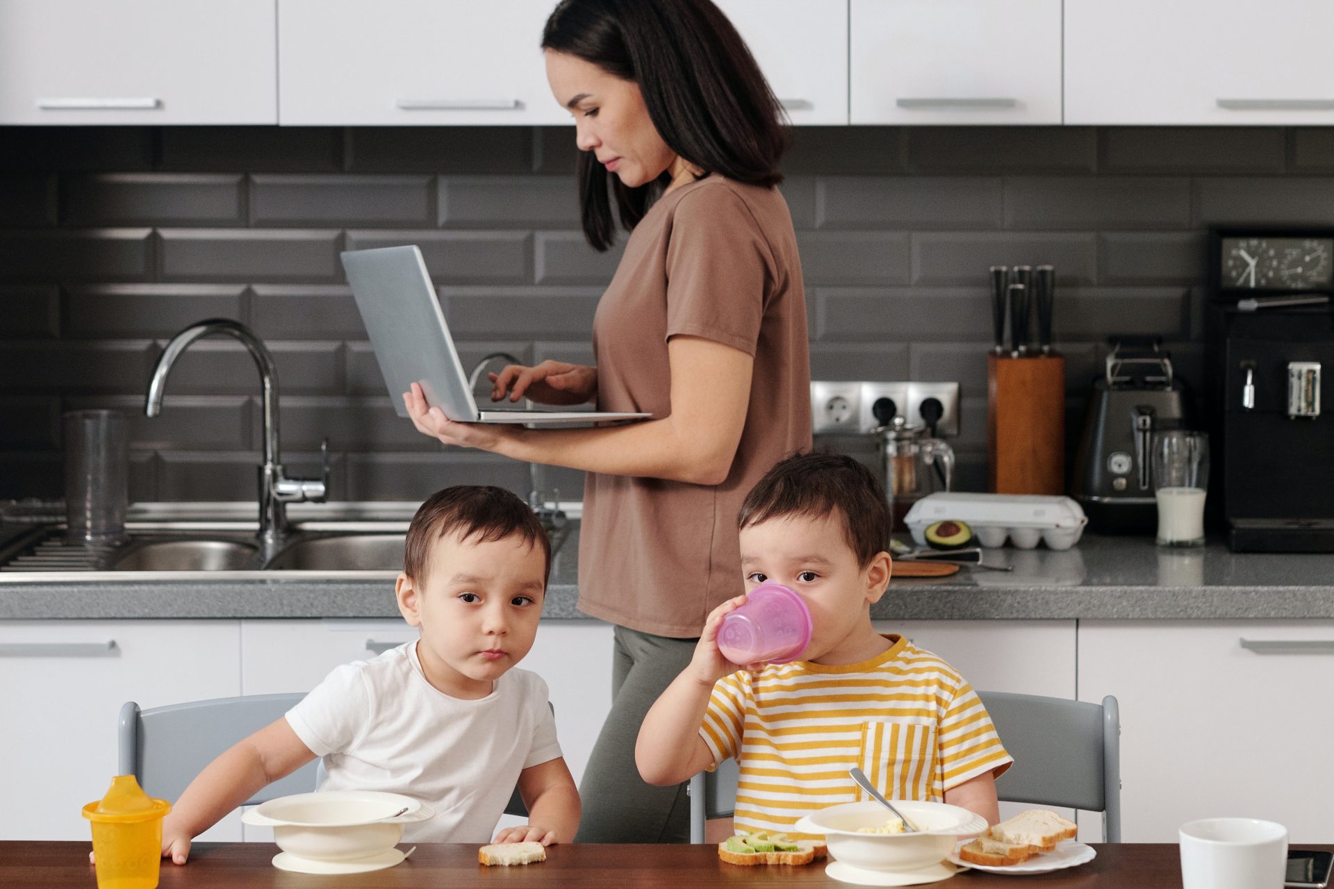 a women working on a laptop behind two children eating at a kitchen table