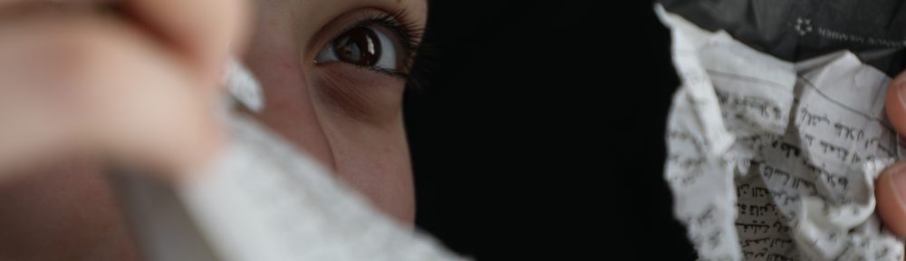 Image of a woman, with her left eye mainly visible, looking though a hole in some torn paper documents.