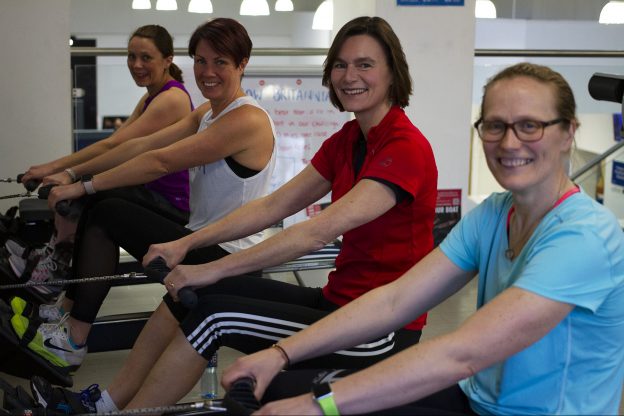 Vice Chancellor Karen Cox and three other female employees all on rowing machines taking part in the Row Britannia challenge, all facing the camera and smiling