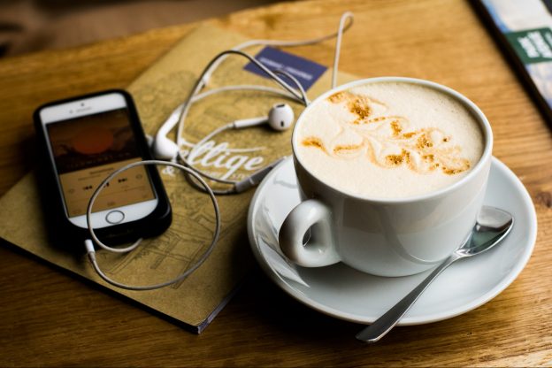 Coffee mug next to a notebook and iphone woth headphones plugged into it