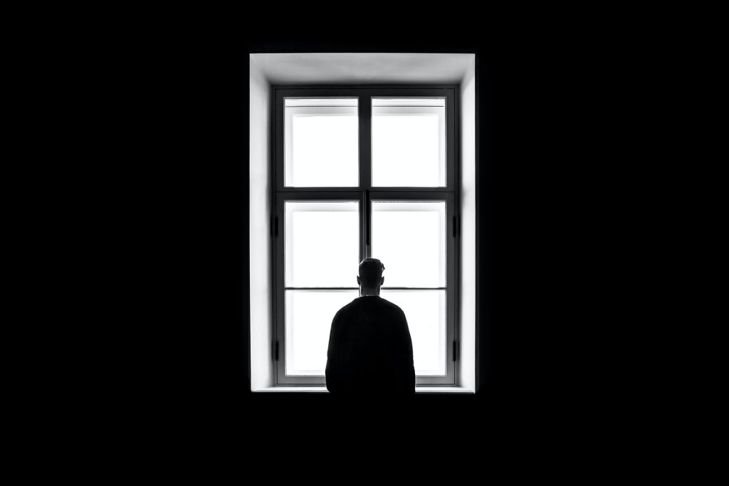 Black and white photo of person standing looking out of window from dark room