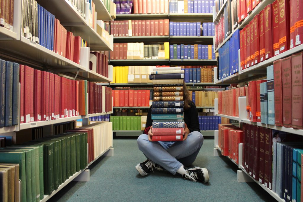 Person sat cross legged in library with pile of books stacked high