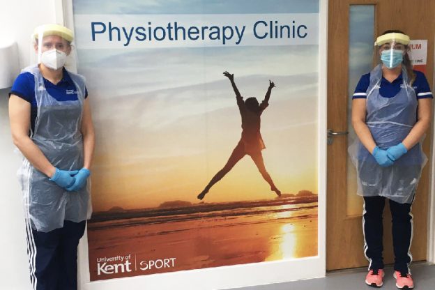 Two Kent Sport staff members in PPE standing next to physiotherapy clinic poster