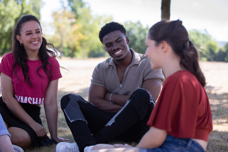photo of 3 young people smiling sitting on the grass, one young white woman in profile in the foreground, a young black man with a short afro and a young white woman with long dark hair facing forwards, all in casual clothes
