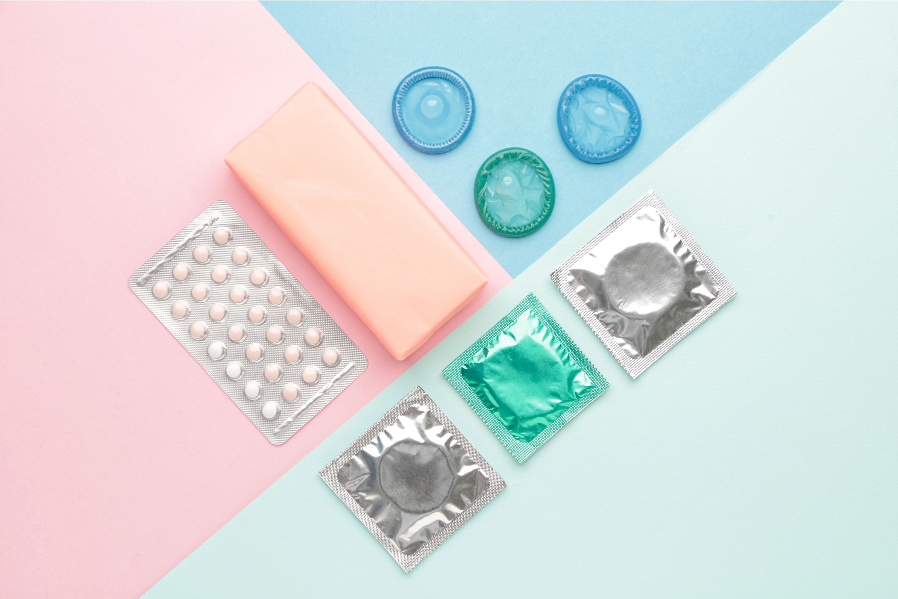 photo of a packet of contraceptive pills, and various condoms, photographed from above on a pastel-coloured background