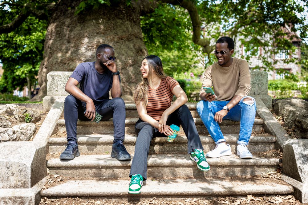 Three students sat laughing together on steps in Westgate Gardens