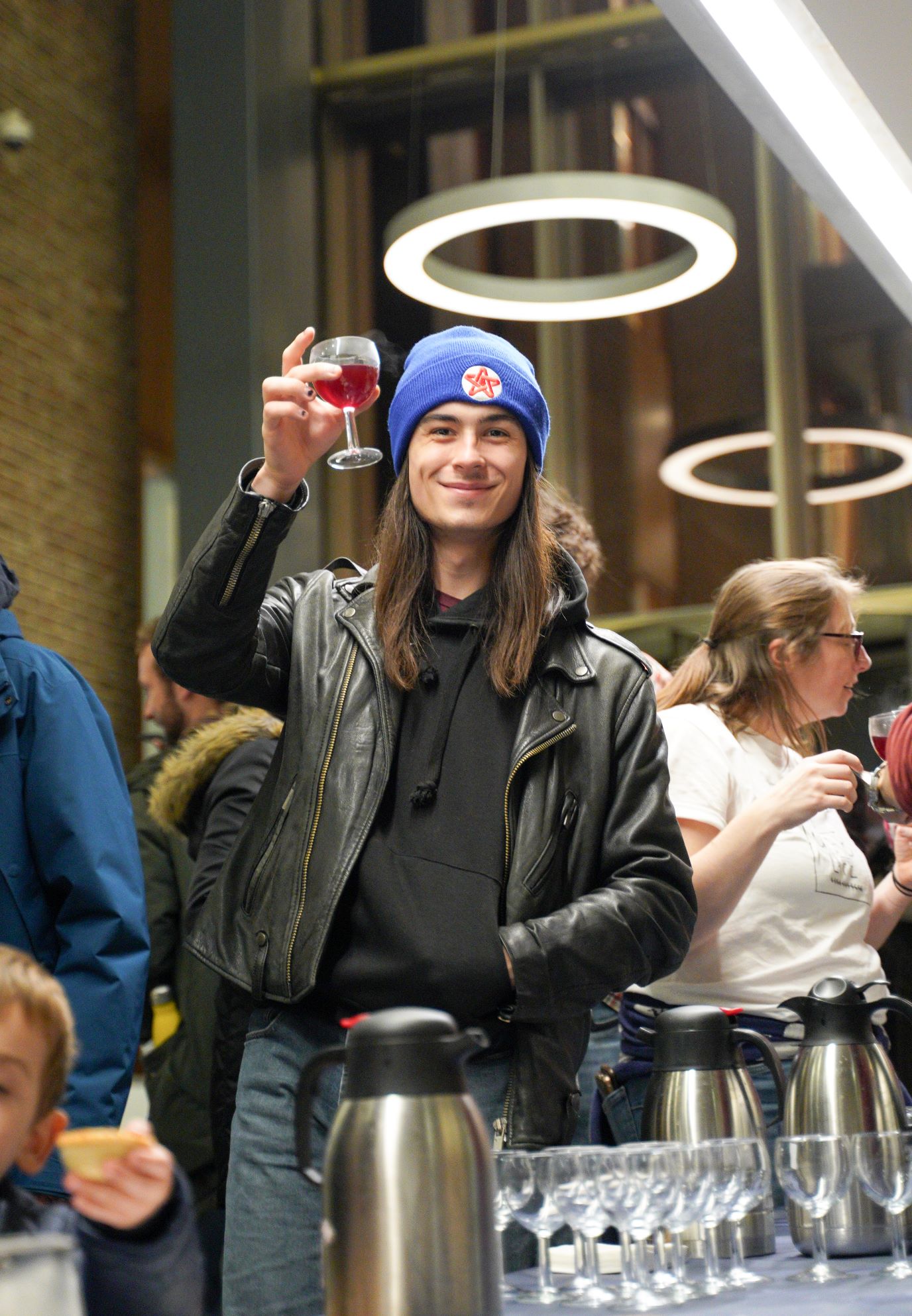 young man with long hair and woolly hat raising his glass and smiling at the camera