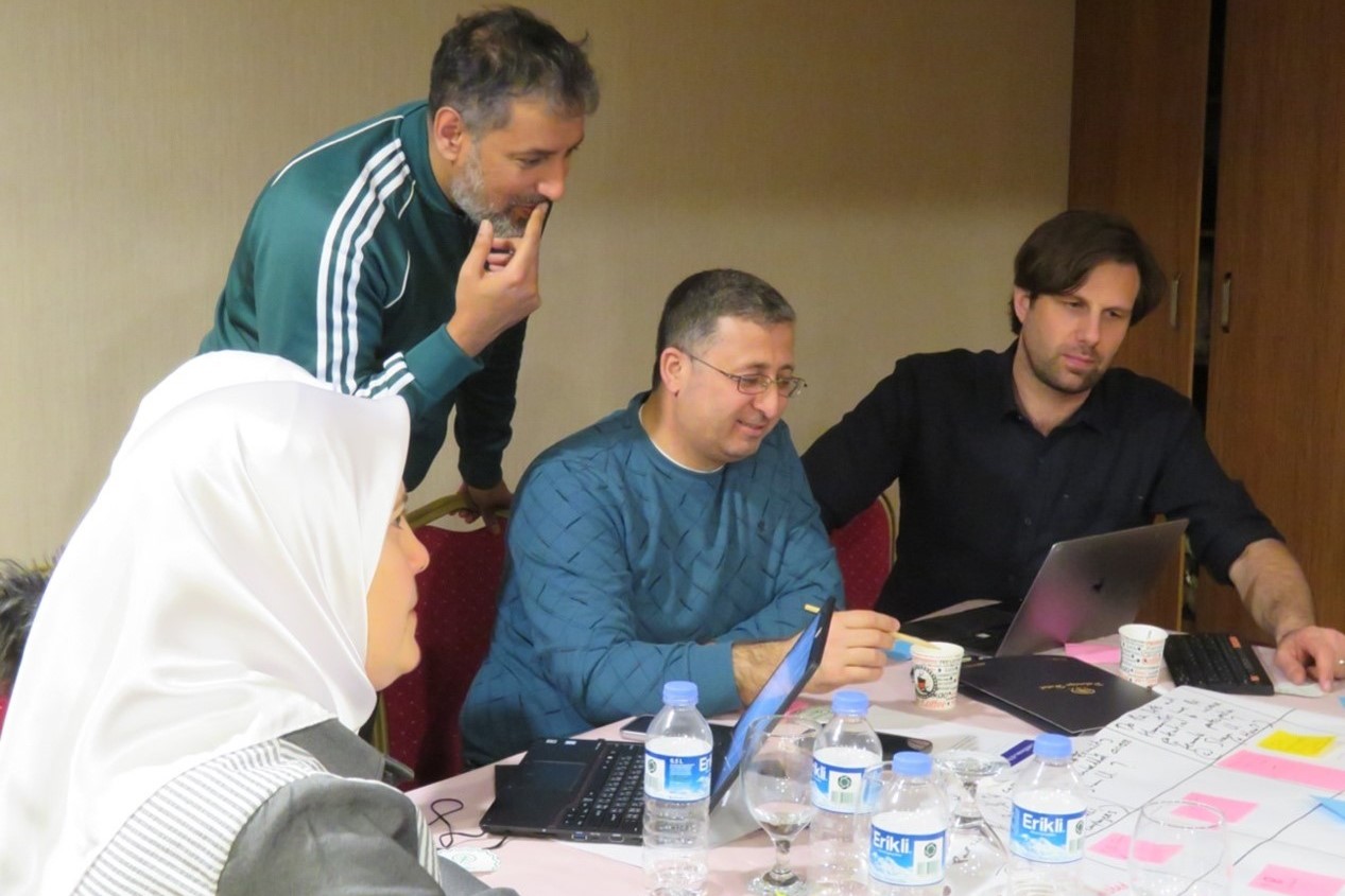 photo of woman in headscarf and three men seated at a table looking at a laptop screen