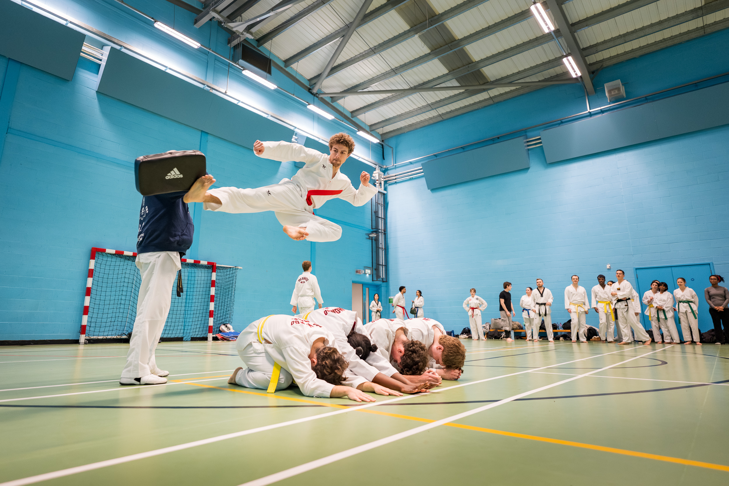 Angus doing a flying side kick over the top of 4 students who are kneeled down on the floor