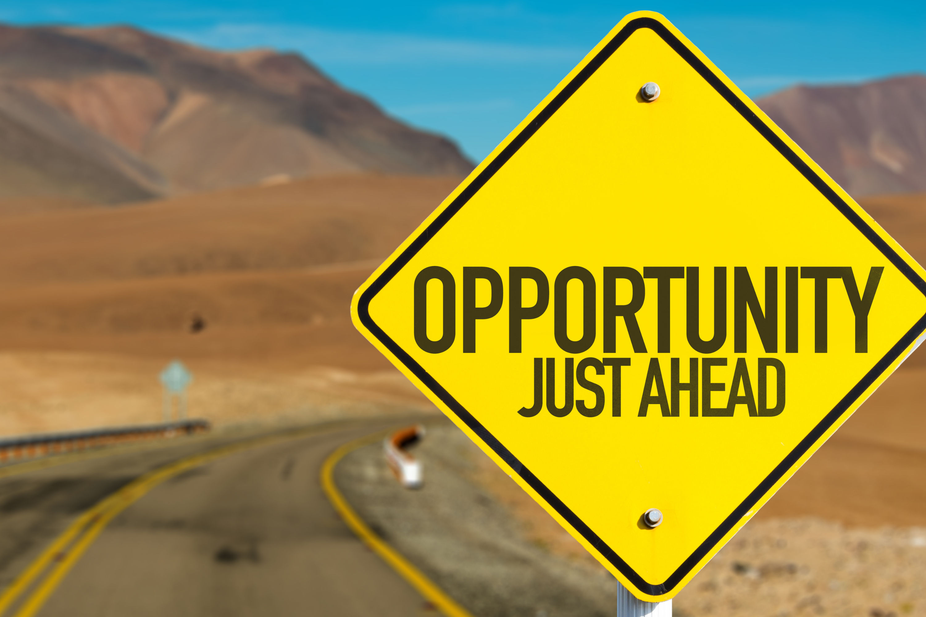 Yellow sign in the forefront reading 'Opportunities Just Ahead' with an open road in the background.
