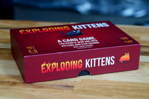Exploding Kittens game box on a wooden table top.